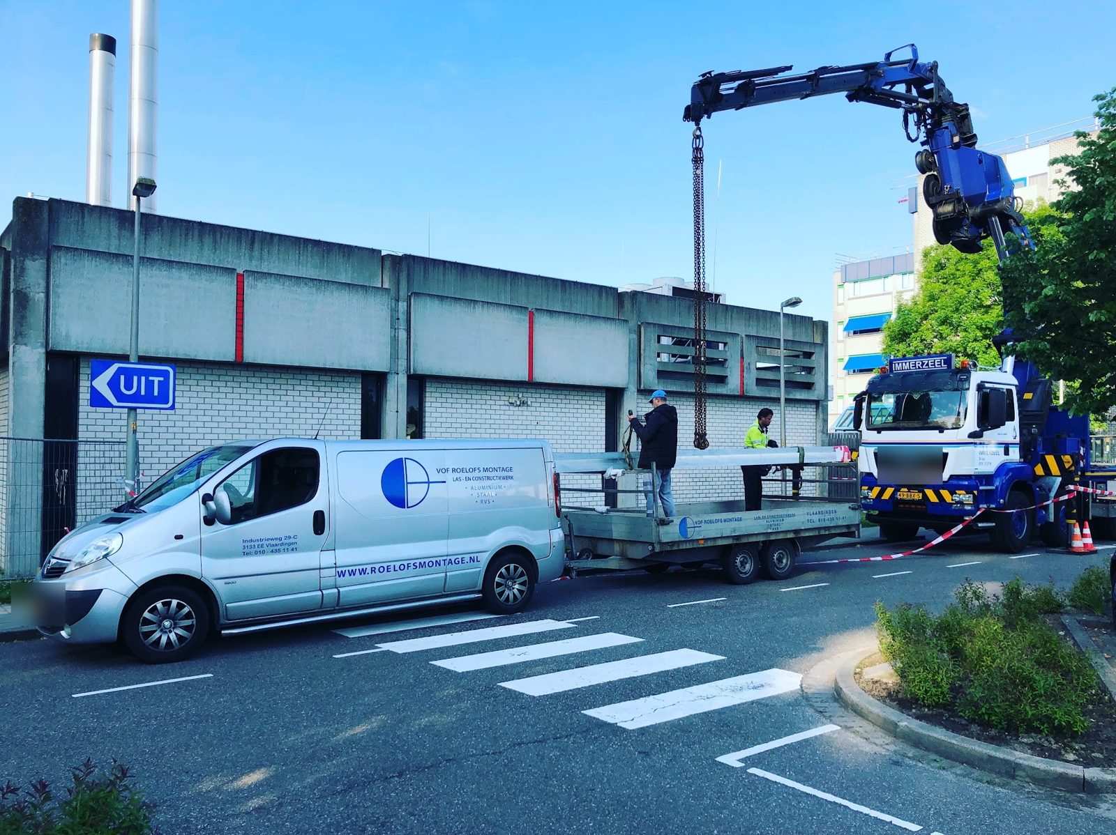Bus en aanhanger van Roelofs Montage bij een gebouw, terwijl een kraanwagen een roestvrijstalen onderdeel hijst